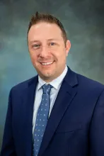 Adam Perkins' headshot. He is wearing a navy suit and smiling at the camera.